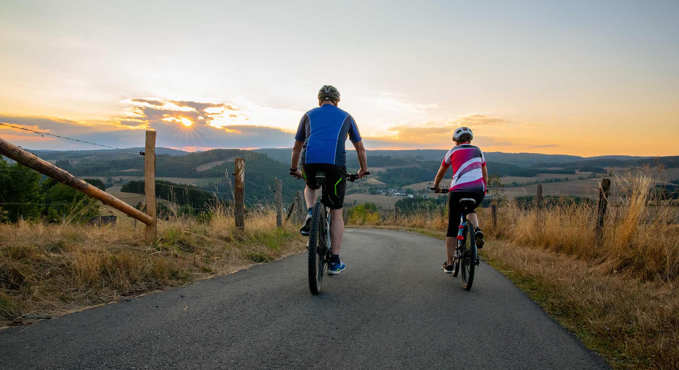 Fahrgemeinschaft: Zwei Radfahrer fahren bei Sonnenuntergang auf einer Straße mit Weitsicht