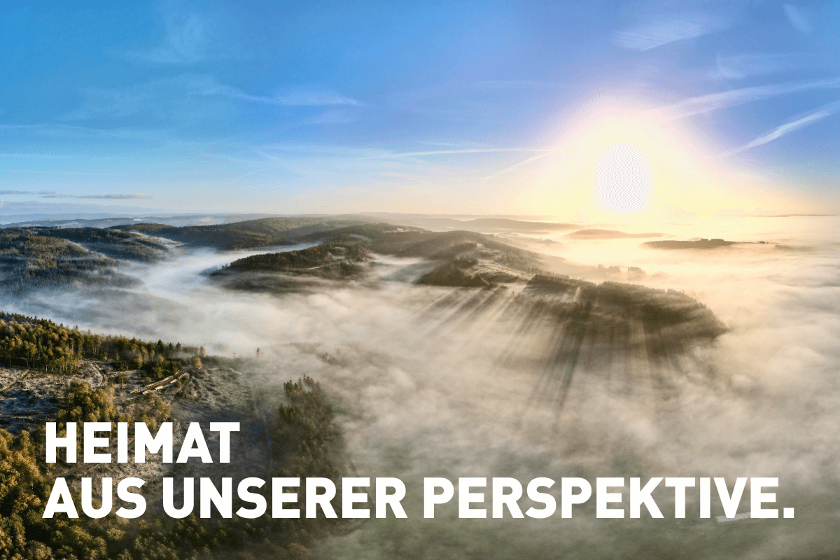 Das Drohnenfoto zeigt ein Panorama der vernebelten Bad Berleburger Wälder über denen die Sonne am blauen Himmel aufgeht. Das Motiv "Heimat" ist das erste der Kampagne "Einfach Ankommen".