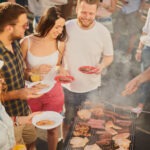 Group of people standing around grill, chatting, drinking and ea